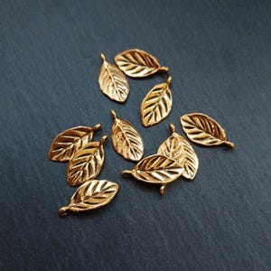 Könnte beinhalten: Eine Ansammlung kleiner, goldfarbener Blattanhänger. Jeder Anhänger hat detaillierte Adern und eine kleine Schlaufe am Stiel. Die Blätter sind auf einer dunkelgrauen Oberfläche verstreut, was eine natürliche und elegante Ästhetik erzeugt. Ideal für Schmuckherstellung oder Bastelprojekte.