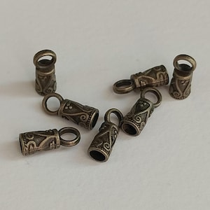 Könnte beinhalten: Nahaufnahme mehrerer antiker, bronzefarbener Metall-Schmuckendkappen mit kleinen Schlaufen. Die Endkappen haben eine zylindrische Form mit dekorativen geätzten Mustern. Diese Schmuckteile werden für die Schmuckherstellung verwendet.