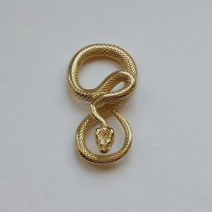Könnte beinhalten: Ein goldfarbener Schlangenanhänger mit detaillierten Schuppen. Die Schlange ist in einer Acht geformt, mit erhobenem Kopf und nach vorne gerichtet. Der Anhänger ist wahrscheinlich aus Metall und hat eine polierte Oberfläche.