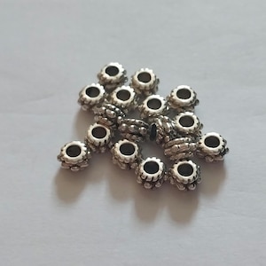 May include: A collection of small, round, silver-toned metal beads featuring a textured, scalloped edge design. Each bead has a central hole and a ridged exterior surface, arranged in a loose cluster on a plain, light-colored surface.