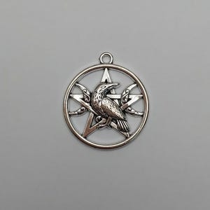 May include: A silver-toned metal pendant featuring a circular frame enclosing a five-pointed star. A detailed raven is positioned in the center of the star, with small crescent moon shapes integrated into the sides of the star points.