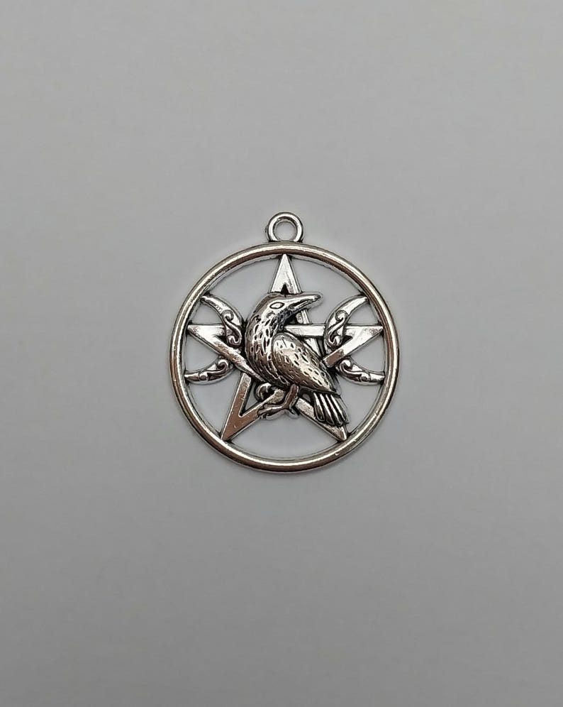 May include: A silver-toned metal pendant featuring a circular frame enclosing a five-pointed star. A detailed raven is positioned in the center of the star, with small crescent moon shapes integrated into the sides of the star points.