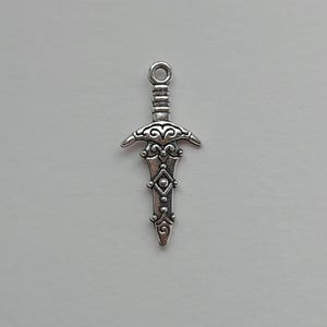 May include: A silver-colored sword charm with intricate detailing. The charm features a decorative hilt and blade, with a small loop at the top for attaching to a necklace or bracelet. The design includes ornate patterns and a pointed tip.