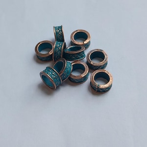 May include: A collection of small, cylindrical metal beads featuring a copper-toned finish with a recessed teal-coloured braided pattern. Each bead has a hollow centre and a textured exterior surface, showing a mix of metallic copper and teal.