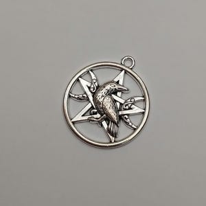 May include: A silver-toned metal pendant featuring a circular frame enclosing a five-pointed star, or pentagram, with a detailed raven perched in the center. Two crescent moon shapes are integrated into the design on either side.
