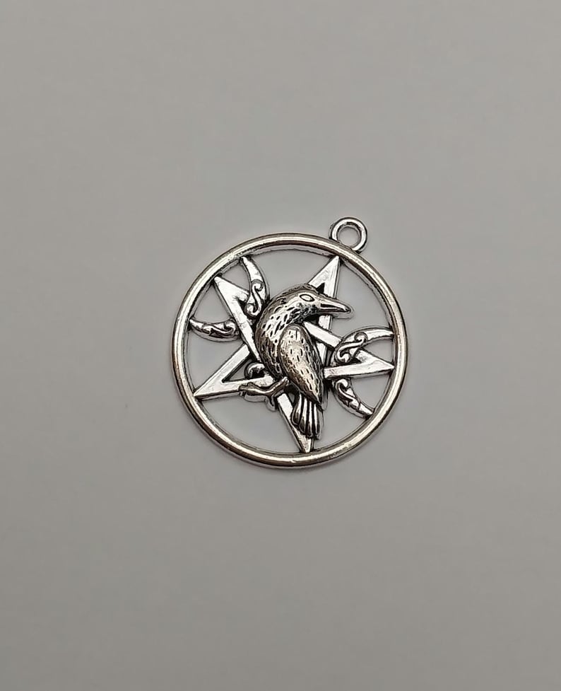 May include: A silver-toned metal pendant featuring a circular frame enclosing a five-pointed star, or pentagram, with a detailed raven perched in the center. Two crescent moon shapes are integrated into the design on either side.