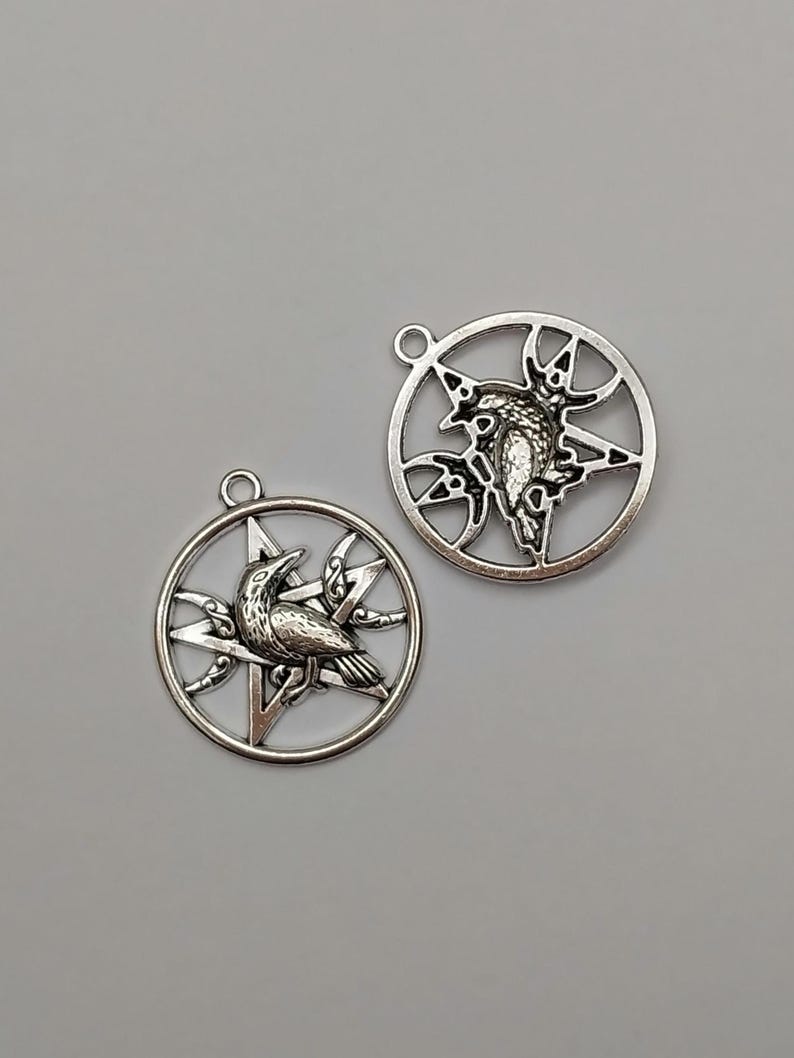 May include: Two silver-toned metal circular pendants, each featuring a five-pointed star design with a central raven figure. Each pendant includes a small loop at the top for attachment, showcasing intricate cut-out details and a metallic finish.