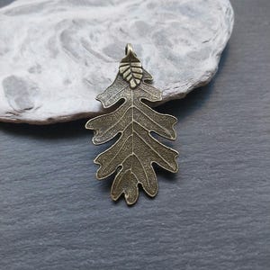 Può includere: Un ciondolo a forma di foglia di quercia in metallo color bronzo con un design dettagliato. La foglia è lunga circa 5 cm e ha una piccola foglia nella parte superiore.