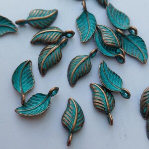 Peut inclure: Une collection de petits charmes en m&eacute;tal en forme de feuille. Chaque charme pr&eacute;sente une surface de couleur sarcelle avec des veines de couleur bronze et une boucle &agrave; la tige. Les charmes sont dispers&eacute;s sur une surface blanche.