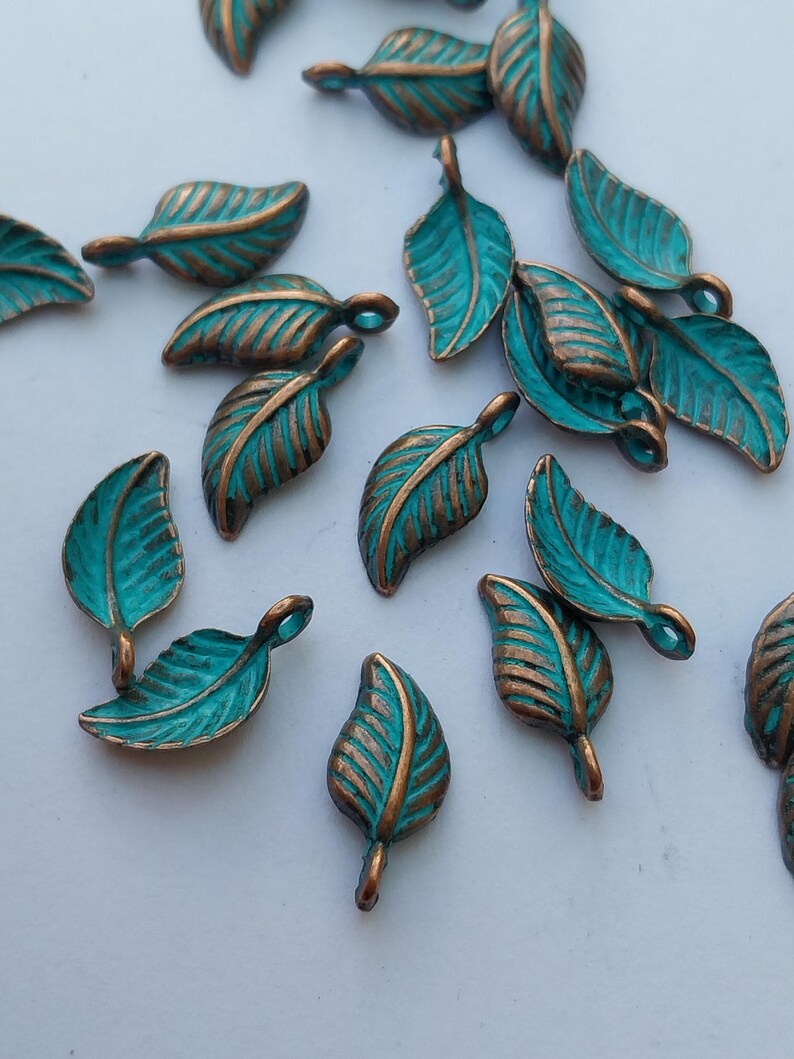 Peut inclure: Une collection de petits charmes en m&eacute;tal en forme de feuille. Chaque charme pr&eacute;sente une surface de couleur sarcelle avec des veines de couleur bronze et une boucle &agrave; la tige. Les charmes sont dispers&eacute;s sur une surface blanche.