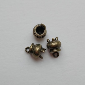 May include: Three small, antique bronze-colored metal charms. Each charm has a unique design, including a small pot shape with a loop for attaching to jewelry or other crafts. The charms are on a neutral background.