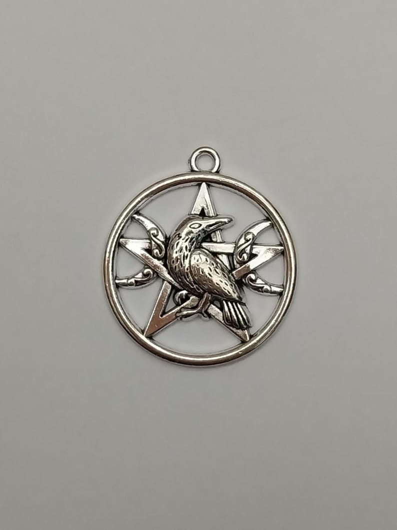 May include: A circular silver-toned metal pendant featuring a central five-pointed star design. A detailed raven is positioned in the center of the star, flanked by two crescent moon shapes on either side. The pendant includes a small top loop.