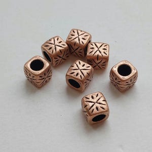 May include: Close-up of seven copper-colored cube beads. Each bead has a dark hole through the center and a carved floral design on each face. The beads are scattered on a light background.