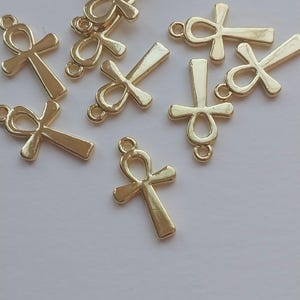 Könnte beinhalten: Eine Sammlung goldfarbener Ankh-Anhänger, verstreut auf hellem Hintergrund. Jeder Anhänger hat die ikonische kreuzartige Form mit einer Schlaufe oben, für Schmuckherstellung oder Basteln. Die Anhänger sind klein und detailliert.