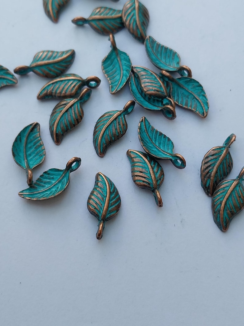 Peut inclure: Plusieurs petits charmes en forme de feuilles de couleur turquoise, avec des veines et des bords de couleur bronze. Ces breloques en m&eacute;tal sont dispers&eacute;es sur une surface blanche, mettant en valeur leur design d&eacute;taill&eacute; et leur finition rustique. Id&eacute;al pour le bricolage.