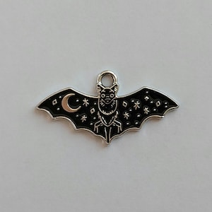 May include: A silver-colored bat charm with black enamel detailing. The bat has outstretched wings with a crescent moon and star accents. The bat's body features a stylized face and decorative elements. A small loop is at the top for attaching to a necklace or bracelet.