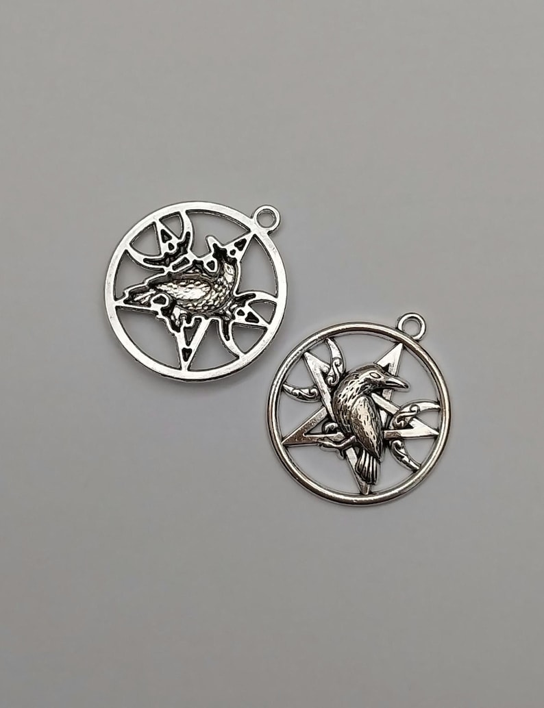 May include: Two silver-toned metal circular pendants, each featuring a five-pointed star design enclosed within a ring. A detailed raven figure is positioned in the center of each star, and each pendant includes a small loop at the top for attachment.