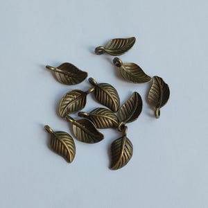 May include: A scattering of small, bronze-coloured metal leaf charms on a white surface. Each leaf charm has detailed veining and a small loop at the top for attaching to jewellery or other crafts. The leaves are a warm, antique gold colour.