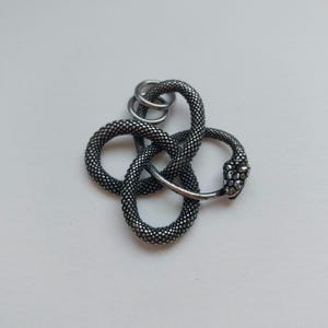 May include: A silver-toned snake pendant with a textured, scale-like design. The snake's head is detailed, and the body is intertwined with three small rings at the top. The pendant is set against a white background.
