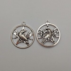 May include: Two circular silver-toned metal pendants, each featuring a central five-pointed star design. Inside the star, a detailed raven is depicted in profile. The star is enclosed within a circular frame, with crescent moon shapes integrated into the design on either side of the raven.