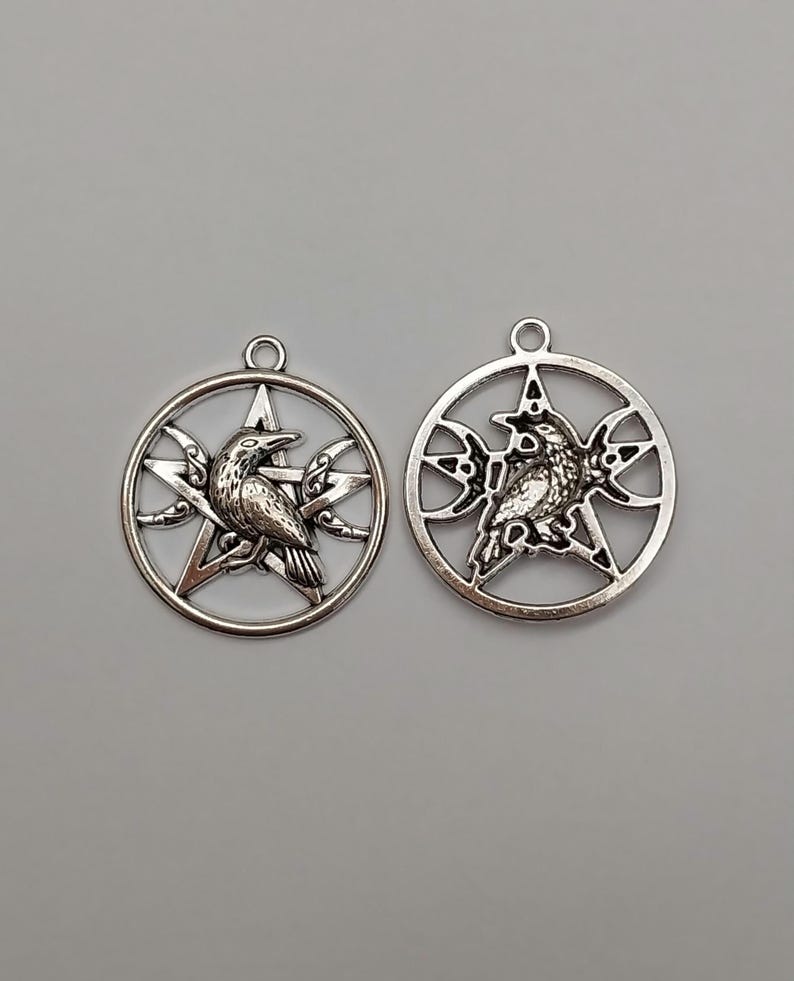 May include: Two circular silver-toned metal pendants, each featuring a central five-pointed star design. Inside the star, a detailed raven is depicted in profile. The star is enclosed within a circular frame, with crescent moon shapes integrated into the design on either side of the raven.