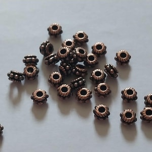 Peut inclure: Une collection de petites perles rondes en métal de couleur cuivre, dotées d'une surface texturée et striée avec un trou central. Les perles sont dispersées dans le cadre, présentant une finition métallique avec des détails sombres en creux.
