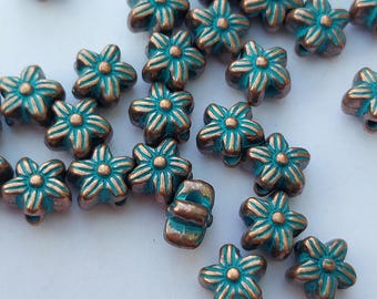 May include: Close-up of numerous flower-shaped beads. The beads are a mix of teal and bronze, with the teal color filling the flower petals and the bronze outlining the petals. The beads are scattered across a white surface.
