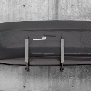 Könnte beinhalten: Schwarze Dachbox mit Carbonfaser-Muster und silbernem Logo. Die Box ist mit zwei schwarzen Halterungen an einer Wand montiert.