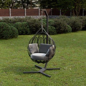 Könnte beinhalten: Ein schwarzer, tropfenförmiger Hängesessel mit grauen Kissen und Polstern. Der Stuhl hängt an einem Metallrahmen und steht auf einem kreuzförmigen Fuß, der auf einem Rasen steht. Im Hintergrund befinden sich grüne Büsche und ein Holzzaun.