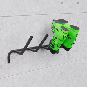 Könnte beinhalten: Schwarzer Ski-Schuhhalter aus Metall, der an einer Betonwand montiert ist, mit zwei Paar grünen Skischuhen, die daran hängen. Die Schuhe haben schwarze Akzente und den Text "FISCHER" und "130" aufgedruckt.