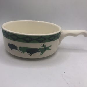 May include: Cream-colored ceramic soup bowl with a handle. The bowl features a green and black geometric border and a repeating graphic of bison and a rider. The handle is white and curved.