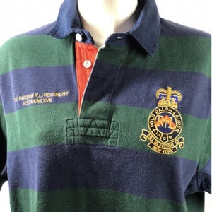 Puede incluir: Una camiseta de rugby a rayas verdes y azul marino con un logotipo de Polo Ralph Lauren bordado en el pecho. El logotipo presenta una corona, un escudo y el texto "Polo Ralph Lauren Bleecker St. New York". La camiseta también tiene el texto "1st Division R.L. Regiment Est. MCMXLXVII" en la manga izquierda.