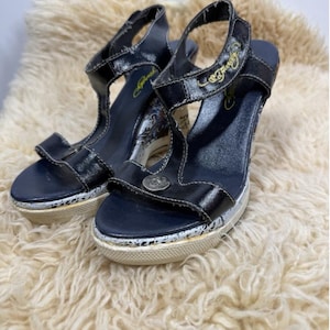 May include: Pair of black platform sandals with T-straps. The sandals have a white sole with a speckled design and a silver emblem on the front. The straps have gold lettering. The shoes are on a fluffy, cream-colored surface.