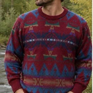 May include: A long-sleeve crewneck sweater featuring a deep red base color with a repeating geometric pattern in shades of blue, teal, and tan. The knit garment has a ribbed collar, cuffs, and hem, paired with a tan baseball cap and blue jeans.