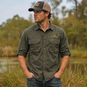 May include: An olive green button-down shirt with four front pockets and a small logo. The person is wearing a black and brown baseball cap. The shirt has rolled-up sleeves and is paired with blue jeans.