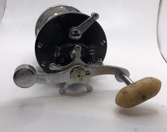 May include: A vintage silver fishing reel with a black spool and a cream-colored handle. The reel has a chrome finish and a simple design.