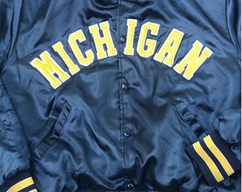 May include: Navy blue satin jacket with the word "MICHIGAN" in yellow block letters across the chest. The jacket has yellow and navy striped cuffs and collar trim. The jacket has a snap front closure.