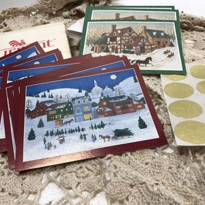 May include: A stack of red and white Christmas cards with a winter village scene. The cards feature a snowy village with houses, trees, and a horse-drawn carriage. The cards are decorated with gold foil accents.