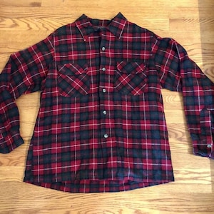 Puede incluir: Una camisa de franela a cuadros rojos, negros y blancos con dos bolsillos en el pecho. La camisa abotonada de manga larga está hecha de una tela suave y tejida. La camisa está tendida sobre una superficie de madera.