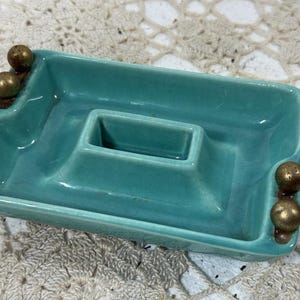 May include: A rectangular, turquoise ceramic ashtray with a central square holder and two sets of brass-colored ball accents on the sides. The ashtray has a glossy finish and a vintage aesthetic.