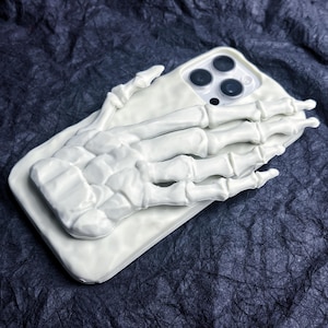 May include: A white phone case featuring a 3D skeleton hand design. The case has a rectangular shape with rounded corners and a camera cutout. The skeleton hand is positioned over the back of the phone case. The background is a dark, textured surface.
