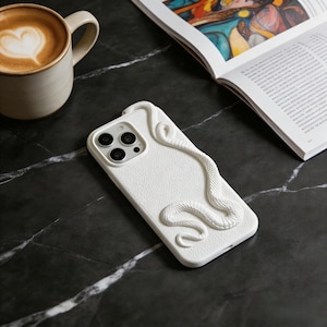 May include: White iPhone case with a textured surface and a raised, sculpted snake design. The case has a camera cutout and is displayed on a black marble surface. A cup of coffee and an open book are also in the image.