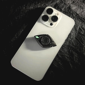 May include: A white smartphone with a black and silver phone grip attached. The phone grip is shaped like an eye and has a glowing green accent. The phone has a triple-lens camera.