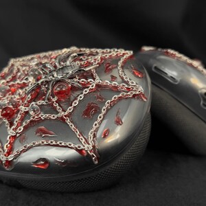 May include: A pair of black wireless headphones with a spiderweb design. The headphones are decorated with silver chains and red beads.