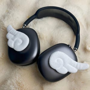 May include: Dark grey over-ear headphones with white felt wing accents. The headphones have a black headband and silver accents. The headphones are resting on a fluffy, cream-coloured surface.