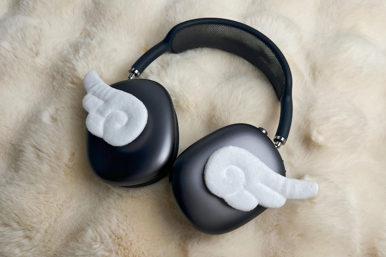 May include: Dark grey over-ear headphones with white felt wing accents. The headphones have a black headband and silver accents. The headphones are resting on a fluffy, cream-coloured surface.