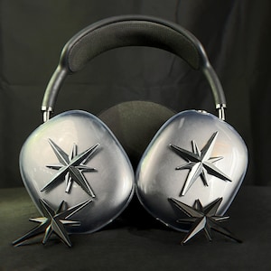 K&ouml;nnte beinhalten: Ein Paar schwarze Over-Ear-Kopfh&ouml;rer mit einem silbernen Sternendesign auf jedem Ohrpolster. Die Kopfh&ouml;rer liegen auf einer schwarzen Oberfl&auml;che.