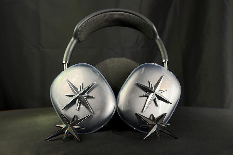 K&ouml;nnte beinhalten: Ein Paar schwarze Over-Ear-Kopfh&ouml;rer mit einem silbernen Sternendesign auf jedem Ohrpolster. Die Kopfh&ouml;rer liegen auf einer schwarzen Oberfl&auml;che.