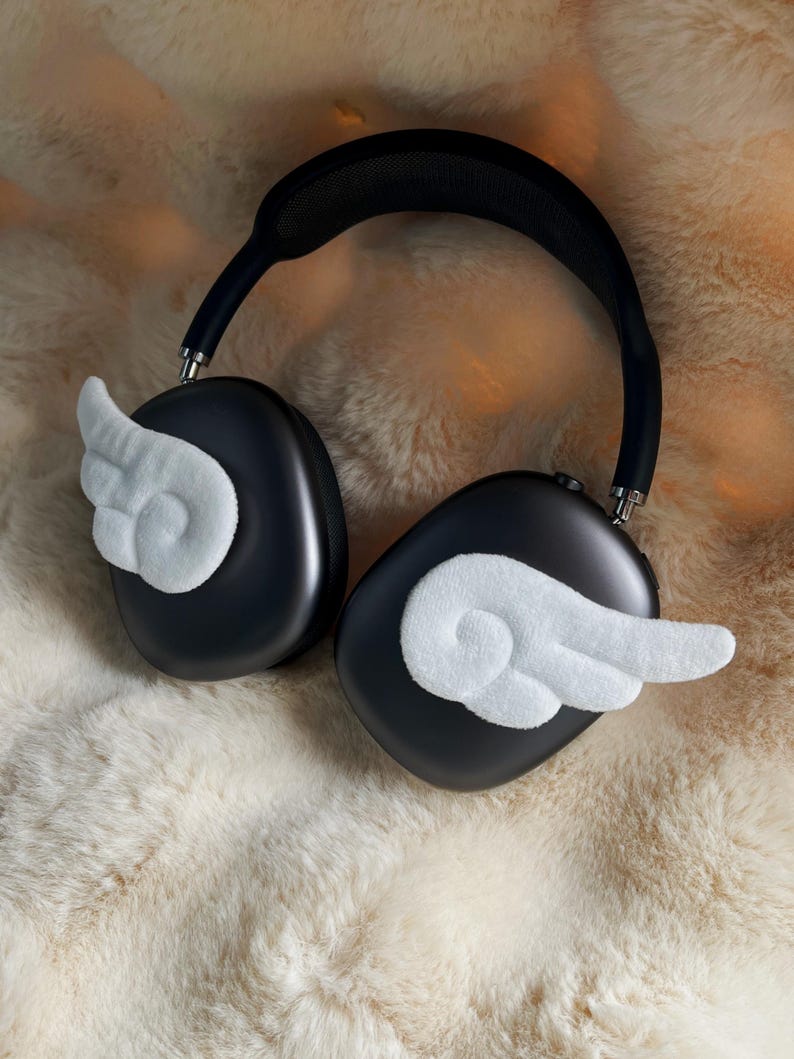 May include: Black over-ear headphones with white, fluffy wing accents. The headphones rest on a cream-coloured, faux fur surface. The earcups are oval-shaped and the headband is black.