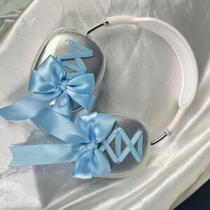 Puede incluir: Un par de auriculares blancos con lazos azules claros en cada auricular. Los lazos est&aacute;n hechos de cinta de sat&eacute;n y est&aacute;n atados en un nudo.